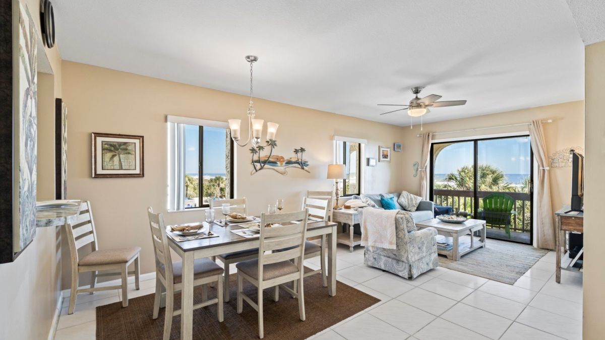 Bright open-plan living and dining area with a coastal view through sliding glass doors, beige walls, and white furniture.