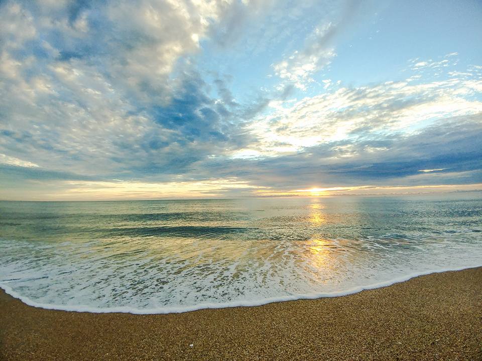 Sunset over a calm sea with gentle waves washing onto a pebbled beach and a partly cloudy sky.
